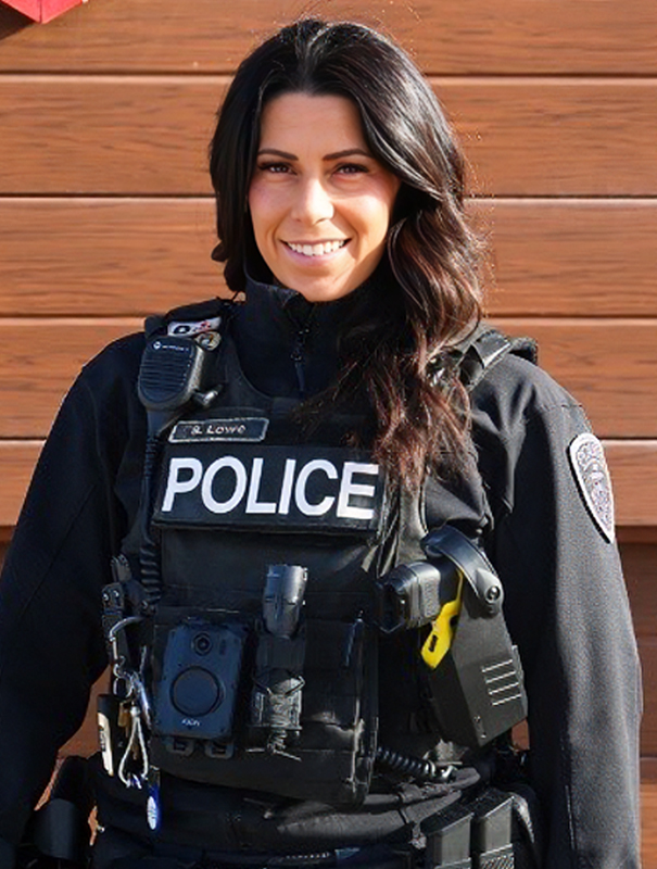 Female police officer wearing a bulletproof vest labeled POLICE, with radio and gear, name tag Lowe, smiling outdoors against a wooden wall.