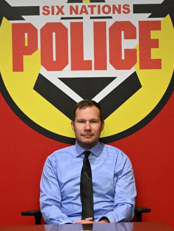 Man in a blue shirt and black tie sits at a desk in front of a large red wall with a yellow-black police emblem reading'SIX NATIONS POLICE'.