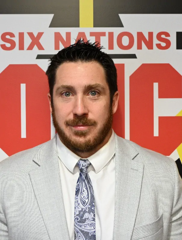 Man in a light gray suit and paisley tie posing in front of a Six Nations banner with red letters.