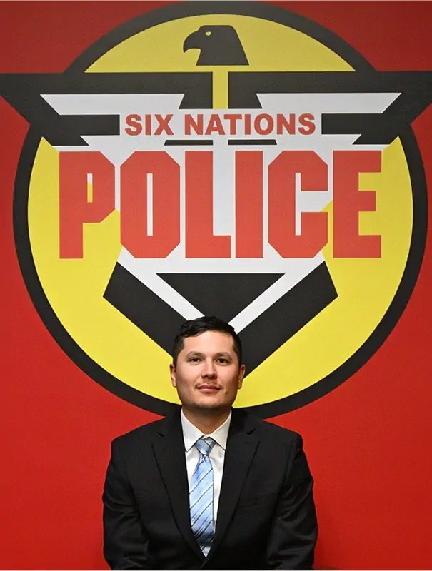 Man in a dark suit and blue striped tie sits in front of a large Six Nations Police emblem on a red wall.