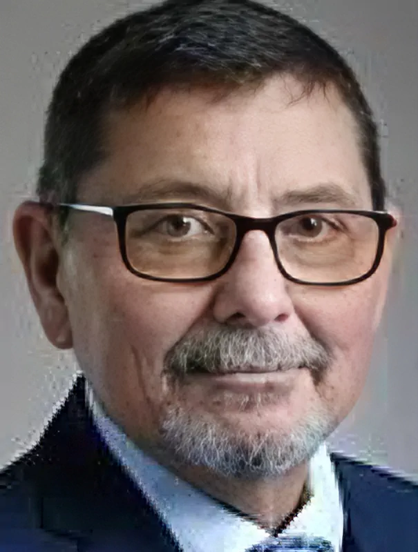 Portrait of a middle-aged man with dark hair, glasses, and a gray beard wearing a dark suit.