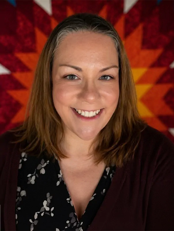 Smiling woman with shoulder-length brown hair in a black floral blouse and maroon cardigan, against a red-orange geometric background.