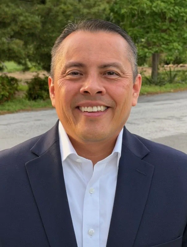 Smiling man in a navy suit and white shirt outdoors, posing for a professional photo.