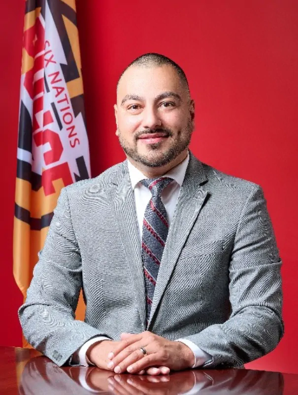 Professional portrait of a man in a gray suit and striped tie, seated at a desk with a colorful flag and red background.